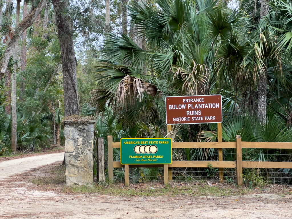 The entry road to Bulow Plantation Ruins Historic State Park in Flagler, Florida. There is a brown sign with the name of the park, an "Amerca's Best State Parks" green sign, a split rail fence, and a lot of lush vegetation.