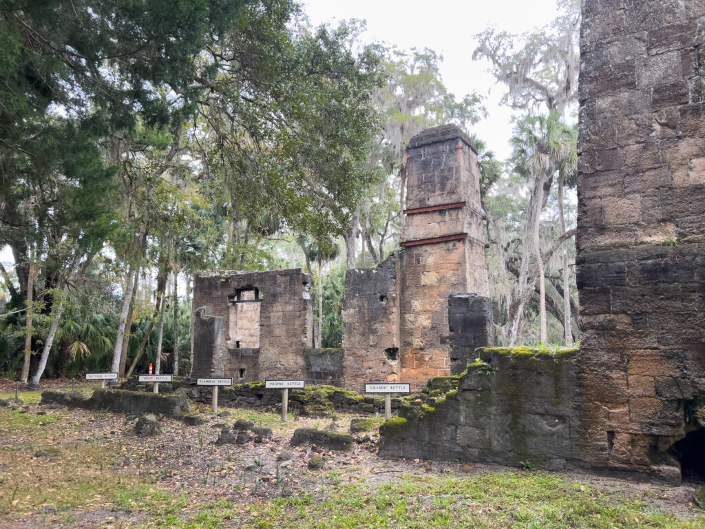 The coquina rock ruins of Bulow Plantation Ruins on a rainy day. The rock stacks are underneath a tree canopy and are pink, gray, and brown. Markers in the ground identify what used to be on the foundations.