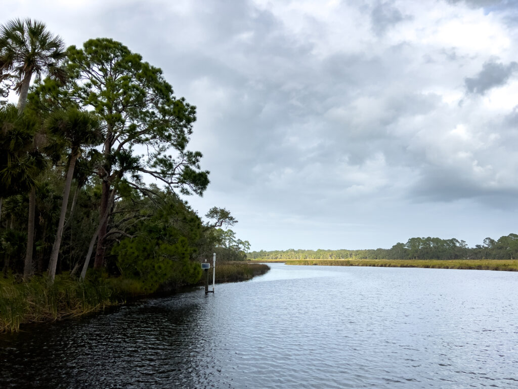 A scenic view of calm and wide Bulow Creek in Bulow Plantation Ruins State Park. One shoreline has tall trees and the other is grasslands and brush.