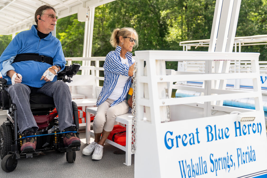 A man in an electric wheelchair wearing a blue shirt sits next to a woman in a blue and white striped collared shirt. They are on the handicap-accessible pontoon boat named "Great Blue Heron" at Wakulla Springs State Park.